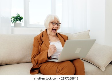 A Very Joyful, Happy Old Lady In A Brown Suit Is Sitting Comfortably On A Beige Sofa Showing Her Fist With Delight And Joy For Success