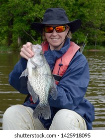 A very large black crappie fish being held vertically by a smiling women angler in a hat and sunglasses on a brown water bay in spring time on a cloudy day