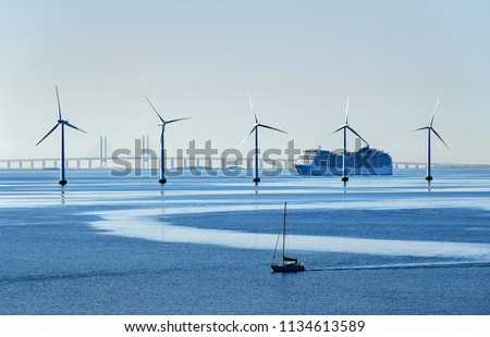 A very large passenger ship and a small sailboat pass offshore wind turbines near the Oresund Bridge between Denmark and Sweden