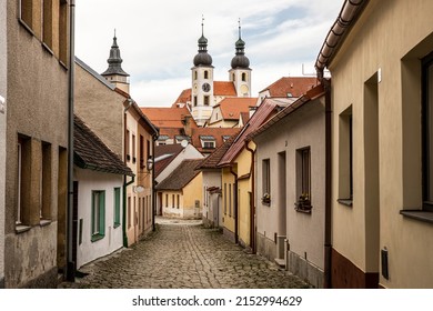 Very narrow street of historical city Telč, Czechia. Cobblestones, small tiny houses, partialy cloudy, big church, towers in the background.