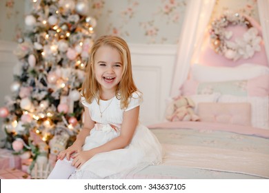 Very nice charming little girl blonde in white dress sitting on a child's bed and laughs loudly on the background of Christmas trees in bright interior of the house