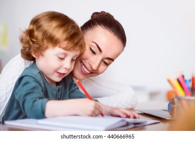 Very nice  Charming smiling mother sitting at the table   teaching her little son drawing