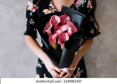 Very nice young woman holding beautiful blossoming mono bouquet of fresh pink calla lilies packed in black wrapping paper on the grey wall background  - Powered by Shutterstock