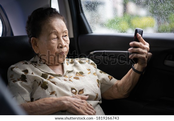 Very old\
Asian passenger woman age between 80 - 90 years old traveling by\
the car while raining and using a video call. Cheerful retired\
woman in a private car portrait with copy\
space.