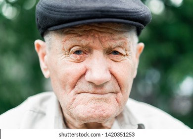 Very Old Caucasian Man Portrait. Grandfather In Hat. Portrait: Aged, Elderly, Loneliness, Senior With Lot Of Wrinkles On Face. Close-up Of A Pensive Old Man Sitting Alone Outdoors.