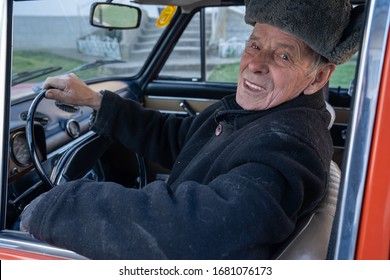 Very Old Confident Man In Black Jacket And Grey Hat Sitting In His Retro Car And Looking At The Camera, Outdoor Photo