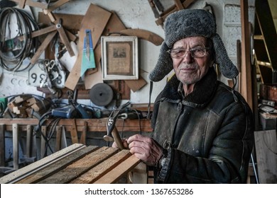 Very Old Man Master In Grey Warm Clothes And Eyeglasses With Hammer In Hands Looking At Camera, Posing Against Wall With Tools