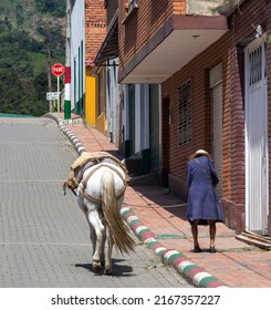 Very old woman walking up a steep street with a white horse that she keeps on a leash with a rope. Villeta, Cundinamarca, Colombia.