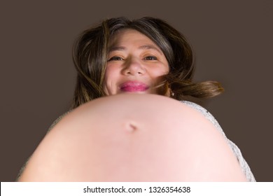 A Very Pregnant Filipino-Caucasian Woman In Her Mid-30s Leans Forward Over Her Baby Bump And Looks Down At The Camera Smiling. Shot From Below With A Clean, Neutral Background.