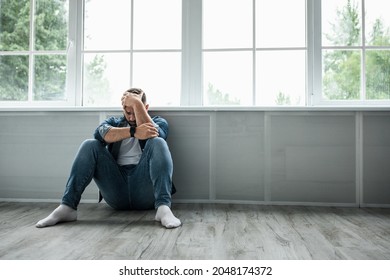 Very sad upset person suffering from grief, loneliness and depression at home, covid-19 lockdown. Unhappy millennial european handsome man sitting on floor near large window and crying, empty space