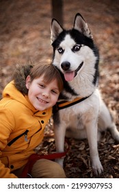 Very Smiling Young European Boy Seven Years Old In The Brown Pants And Yellow Jacket Is Sitting On The Ground With The Husky Dog In The Autumn Park