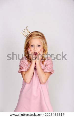 Very surprised little girl in a pink dress. She has a crown on her head. A little girl is playing princess. Child on a light background