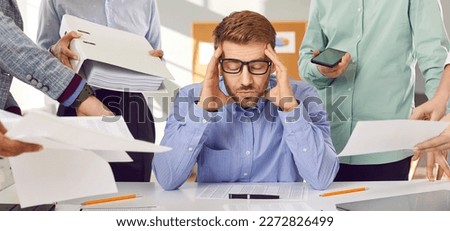 Very tired, stressed and busy man at work. Young man in shirt and glasses sitting at office table, holding his head, and ignoring colleagues giving him lots of business paperwork. Banner background