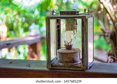 A Very Ugly Old And Dirty Kerosene Lamp On A Wooden Bench With A Blurred Bushland Background