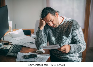 A Very Upset Man In A Warm Sweater Is Sitting At A Table With Utility Bills In His Hands.  A Man Emotionally Reacts To The Increased Costs Of Heating, Gas And Electricity Using A Calculator