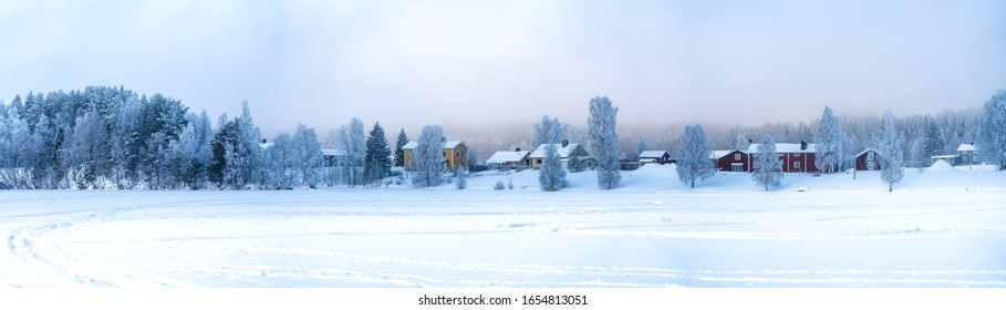 Very wide panorama view on Swedish village with red and yellow wooden houses in winter overcast on frozen river coast at spruce forest edge, Lappland, Scandinavia.