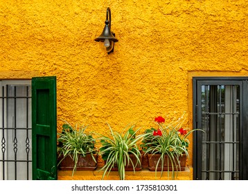 A Very Yellow Rough Wall On An Old House With Flowers And Wall Light Between Two Windows In Italy