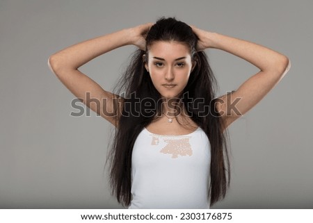 Very young woman in a white tank top gathers her hair, showcasing her beautiful skin and neck. The beauty of youth and a small pendant necklace. Long black hair and intense gaze