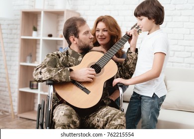 The Veteran In A Wheelchair Came Back From Army. A Man In Uniform In A Wheelchair With His Family. The Veteran Is Playing The Guitar. Wife And Son Listen To His Music.