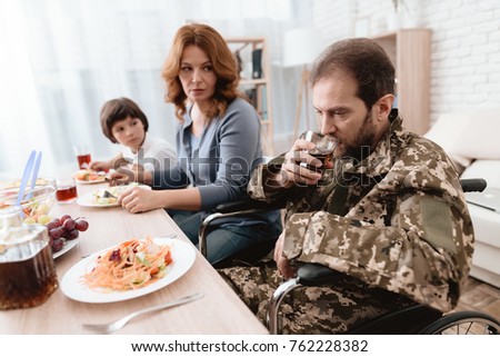 A veteran in a wheelchair dines with his family. A man in military uniform is sitting at the kitchen table. The veteran is depressed. He has problems with alcohol.