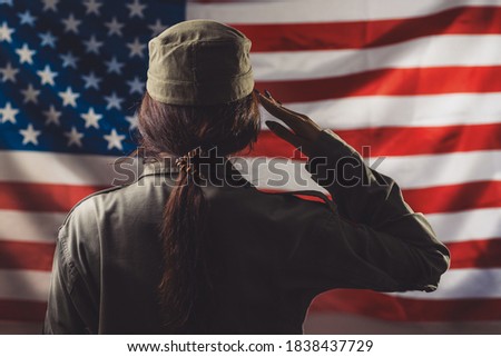 Veterans Day, Memorial Day, Independence Day. A female soldier saluting against the background of the American flag. Rear view. Concept of national American holidays and patriotism