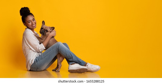 Veterinary Clinic Advertisement Concept. Happy Afro Woman Dog Owner With Her Cute French Bulldog Puppy, Sitting On Floor Over Yellow Background