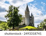 Veves Castle on hill against blue sky, towers with conical roofs and stone walls, a huge tree with green leaves, a sunny day in Houyet, Namur province, Wallonia, Belgium