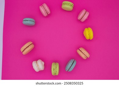 A vibrant arrangement of macarons forming a circular pattern on a bright pink background. The macarons are in various pastel colors, including pink, green, blue, yellow, peach, and white.