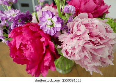 A vibrant arrangement of pink peonies, purple flowers, and green hydrangeas in a clear vase on a wooden table, showcasing a colorful and fresh floral centerpiece indoors. - Powered by Shutterstock