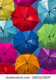 A vibrant array of colorful umbrellas forms a dense, cheerful canopy against a clear blue sky. Sunlight glows through the translucent nylon fabric, emphasizing the crisp radial skeletal structure.
