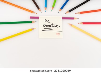 A vibrant array of sharpened colored pencils is arranged in a dynamic semi-circle, all pointing inwards towards a central yellow sticky note. The note, handwritten with the encouraging phrase 'Be creative' and a small red heart, serves as an inspiring focal point for artistic expression and imagination. Against a clean, bright white background, the diverse hues of the pencils create a visually appealing composition, illuminated by soft, even studio lighting. This setup provides ample copy space, perfect for themes of education, innovation, and creativity.