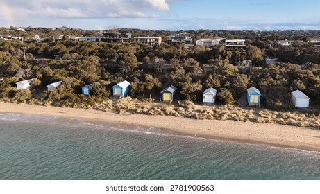 Vibrant bathing boxes sit along a sandy beach with coastal vegetation and distant residential houses.