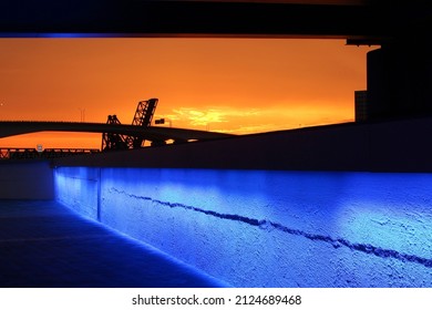 Vibrant blue illuminated stone wall under a bridge in a coastal industrial area at colorful orange sunset.