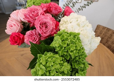 A vibrant bouquet featuring pink roses, red roses, green hydrangeas, and white hydrangeas, accented with eucalyptus leaves, arranged on a wooden table in a cozy indoor setting. - Powered by Shutterstock