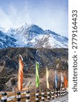 Vibrant Buddhist prayer flags flutter along a mountain road with a dramatic backdrop of towering snow-covered Himalayan peaks in the Annapurna region of Nepal. The rugged alpine landscape