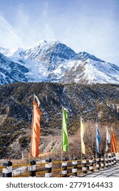 Vibrant Buddhist prayer flags flutter along a mountain road with a dramatic backdrop of towering snow-covered Himalayan peaks in the Annapurna region of Nepal. The rugged alpine landscape