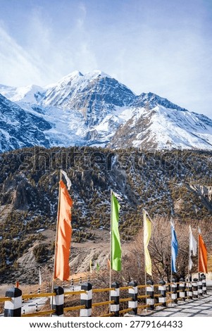 Vibrant Buddhist prayer flags flutter along a mountain road with a dramatic backdrop of towering snow-covered Himalayan peaks in the Annapurna region of Nepal. The rugged alpine landscape