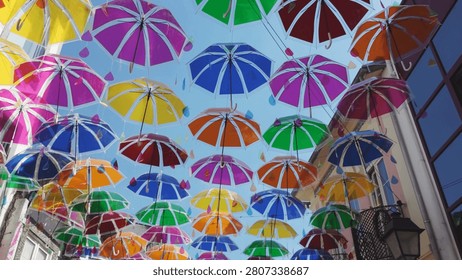 A vibrant canopy of translucent multi-colored umbrellas creates a festive atmosphere in an urban alleyway. The sunlight filters through the fabric, casting bright, saturated hues onto nearby buildings.