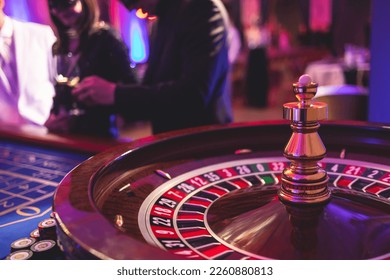 Vibrant casino table with roulette in motion, with casino chips, tokens, the hand of croupier, dollar bill money and a group of gambling rich wealthy people playing bet in the background
