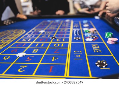 Vibrant casino table with roulette in motion, with casino chips, tokens, the hand of croupier, dollar money and a group of gambling rich people playing bet, blue poker table and deck of cards