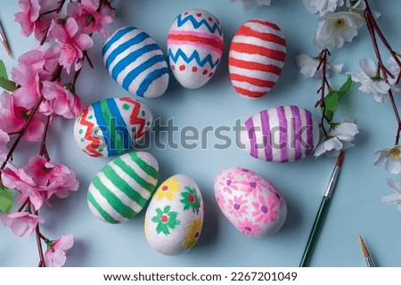 Vibrant and cheerful flat lay background features circle of colorful Easter eggs adorned with festive decorations, surrounded by spring blossoms and brushes. For website, social media, greeting cards