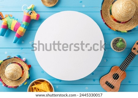 Vibrant Cinco de Mayo setup: top view sombreros, vihuela, cactus, pinata, nachos and confetti on wooden blue backdrop. Empty circle great for themed advertising