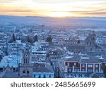 Vibrant cityscape during twilight with traditional architecture and terracotta roofs in the foreground, modern buildings with illuminated windows in the middle ground, a central boulevard with trees a