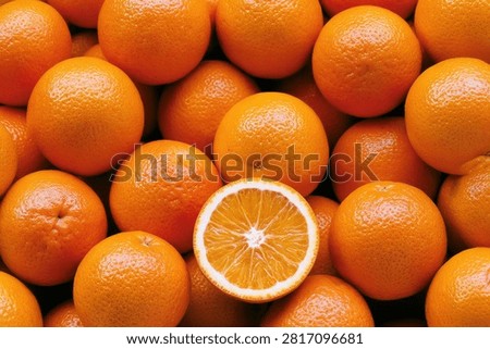 A vibrant, close-up view of a large stack of ripe, whole oranges, with one orange cut in half revealing its juicy interior and seeds.