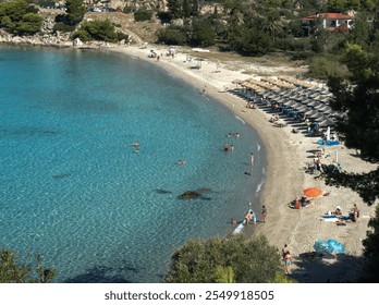 A vibrant coastal scene featuring a sandy, curved beach, clear blue water, and a lively relaxation area with sun loungers and umbrellas. - Powered by Shutterstock