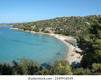 A vibrant coastal scene featuring a sandy, curved beach, clear blue water, and a lively relaxation area with sun loungers and umbrellas. - Powered by Shutterstock