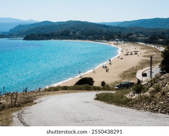 A vibrant coastal scene featuring a sandy, curved beach, clear blue water, and a lively relaxation area with sun loungers and umbrellas. Lush greenery and scenic hillside complete the peaceful beach  - Powered by Shutterstock