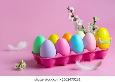 Vibrant collection of painted Easter eggs arranged in a pink egg tray, adorned with delicate flowers and feathers, set against a soft pink background.