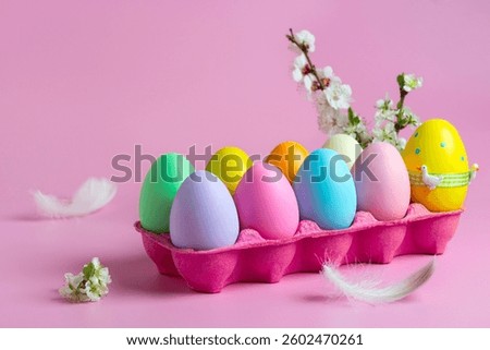 Vibrant collection of painted Easter eggs arranged in a pink egg tray, adorned with delicate flowers and feathers, set against a soft pink background.