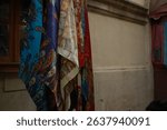 A vibrant display of traditional scarves with intricate paisley and floral patterns, hanging against an old stone wall in a historic district. 